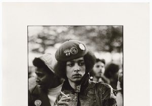 A young black woman with a somber expression wearing a beret with political buttons and a leather jacket. She's holding a disposable coffee cup and a cigarette.
