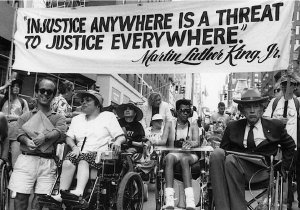Several wheelchair users and a little person marchi in a parade with a banner