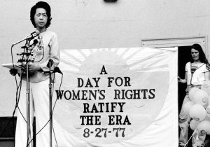 A Chinese American woman speaks into multiple microphones at a rally. A sign behind her reads &quot;A Day for Women's Rights. Ratify the ERA (Equal Rights Amendment) 8-27-77&quot;
