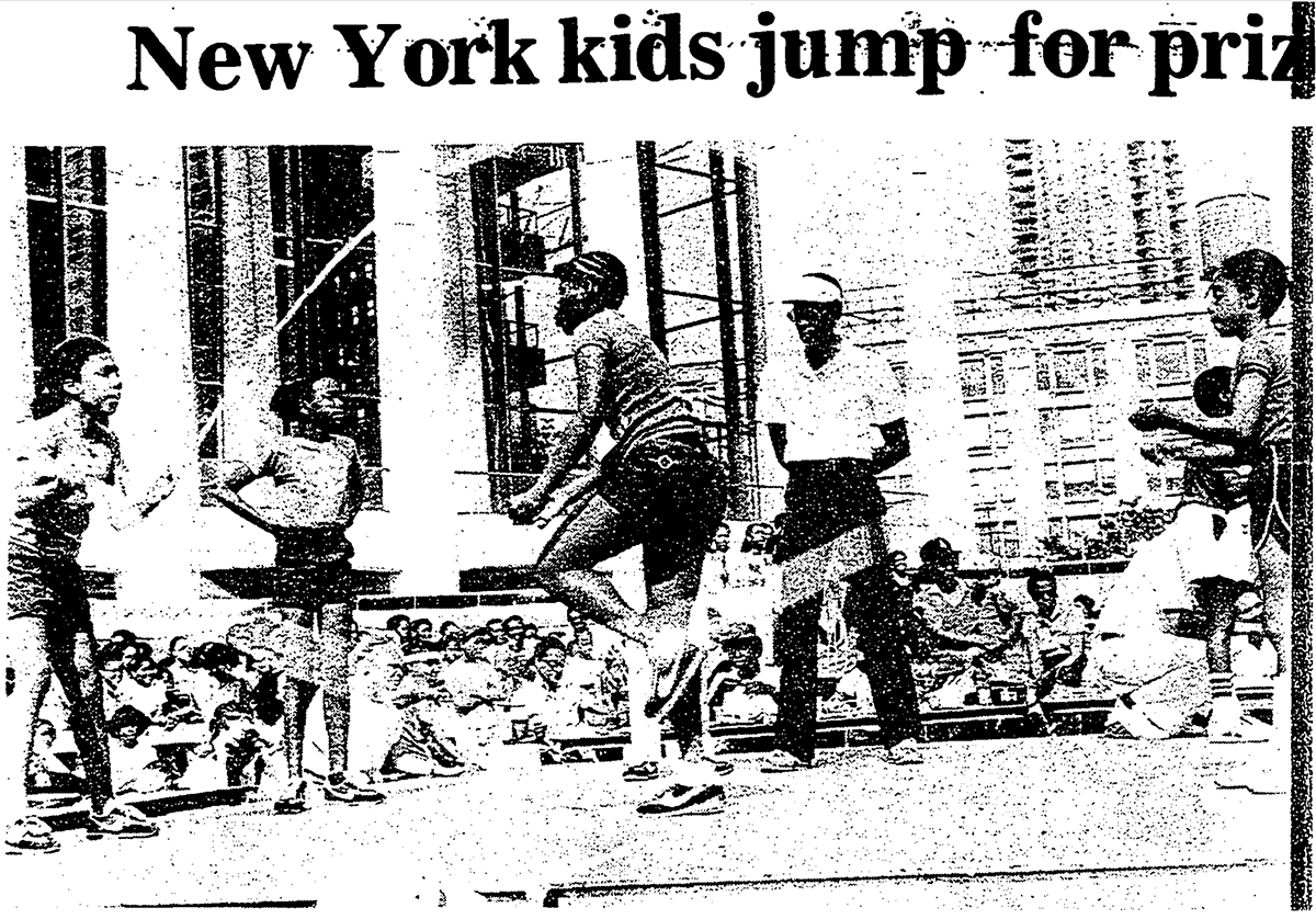 Black children jump rope in front of a crowd of seated people. A referree watches closely.