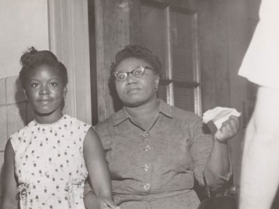 A middle-aged Black woman holds the arm of her teenaged daughter. They stand in a doorway, likely at a school.